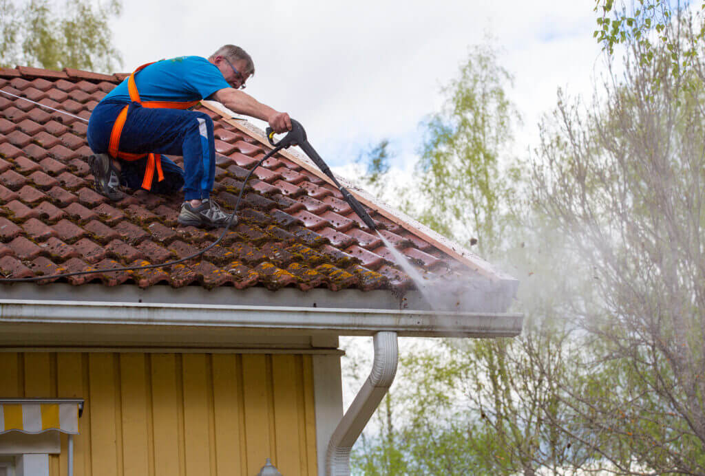 A Caucasian man wearing a safety harness pressure washes a roof and gutters on a slippery surface.