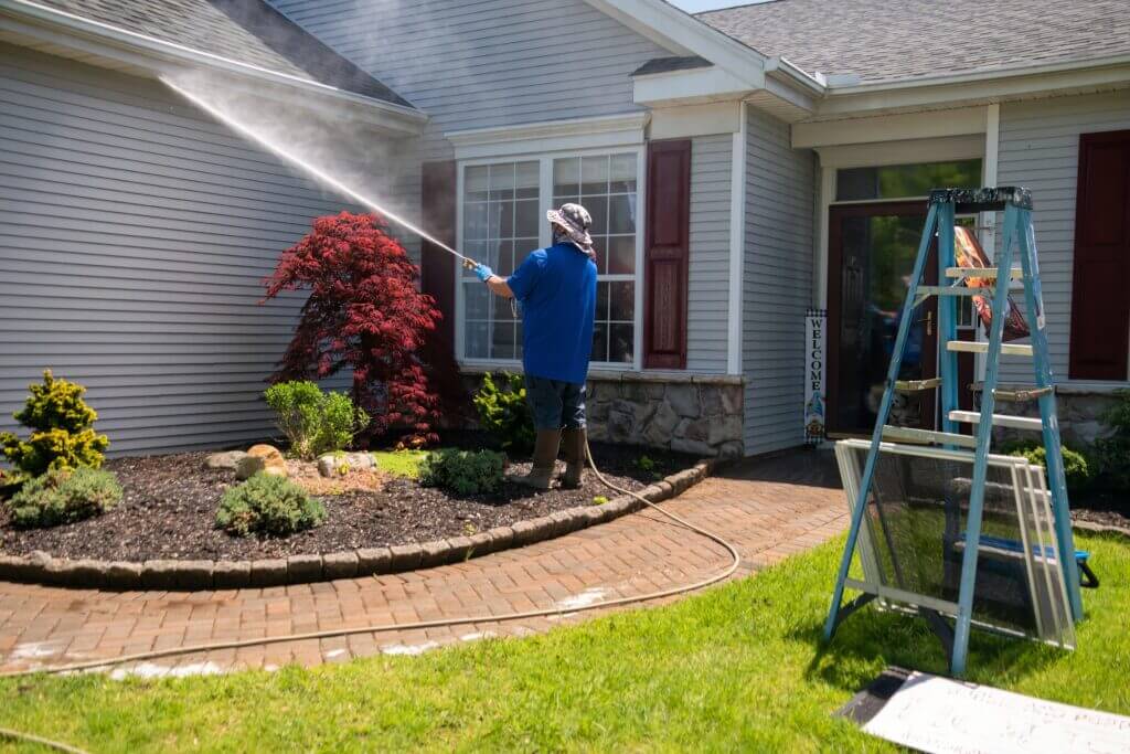 A man using a power washer to clean the underside of a roof and house walls, removing dirt and mold.