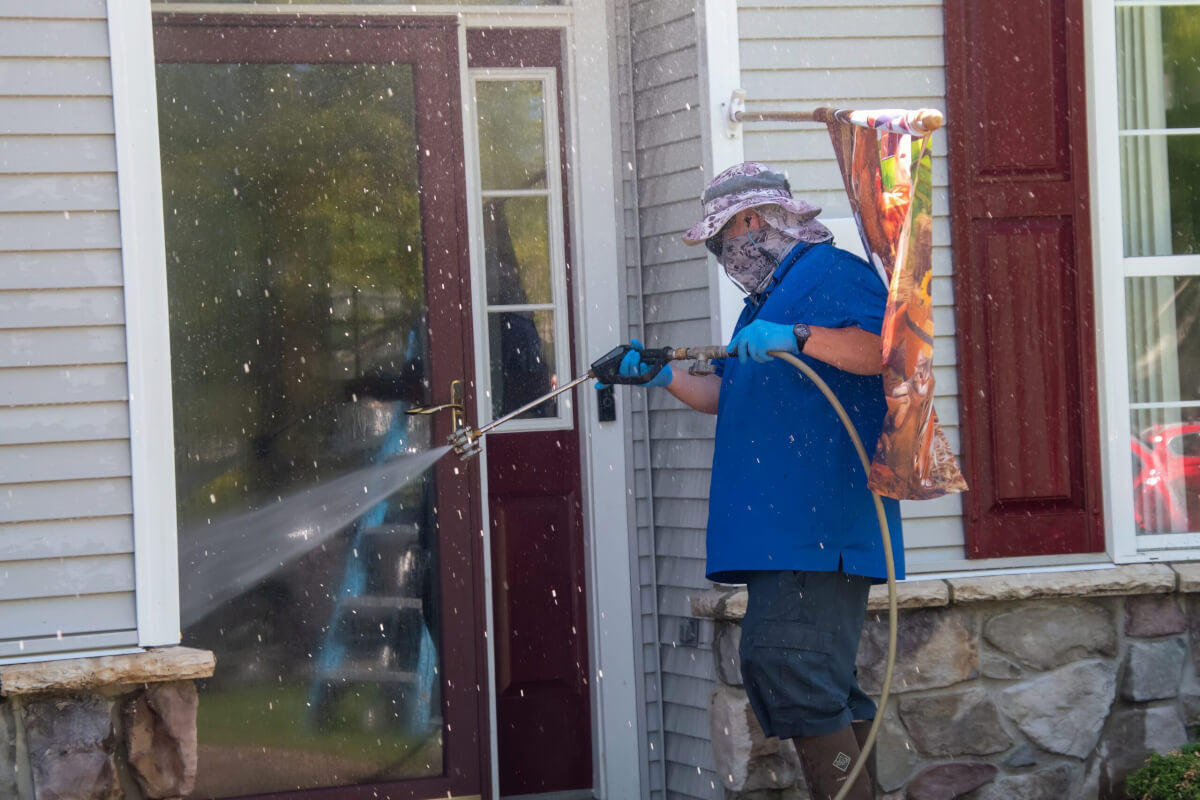 A Caucasian man wearing latex gloves uses a water sprayer wand to power wash the front of a house, removing dirt and grime.