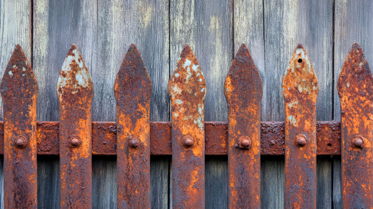A close-up of a heavily rusted metal fence with corroded spikes needing resurfacing.