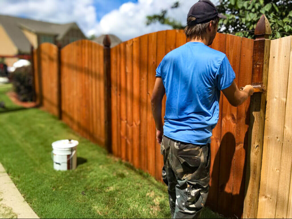Man staining a wooden fence with a brush, applying a protective wood stain and sealant for long-lasting durability and rich color.