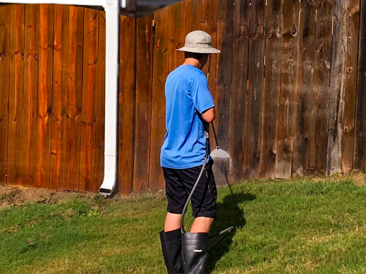 A worker pressure washing a wooden fence to remove dirt, grime, and old stain before applying a protective stain and seal.