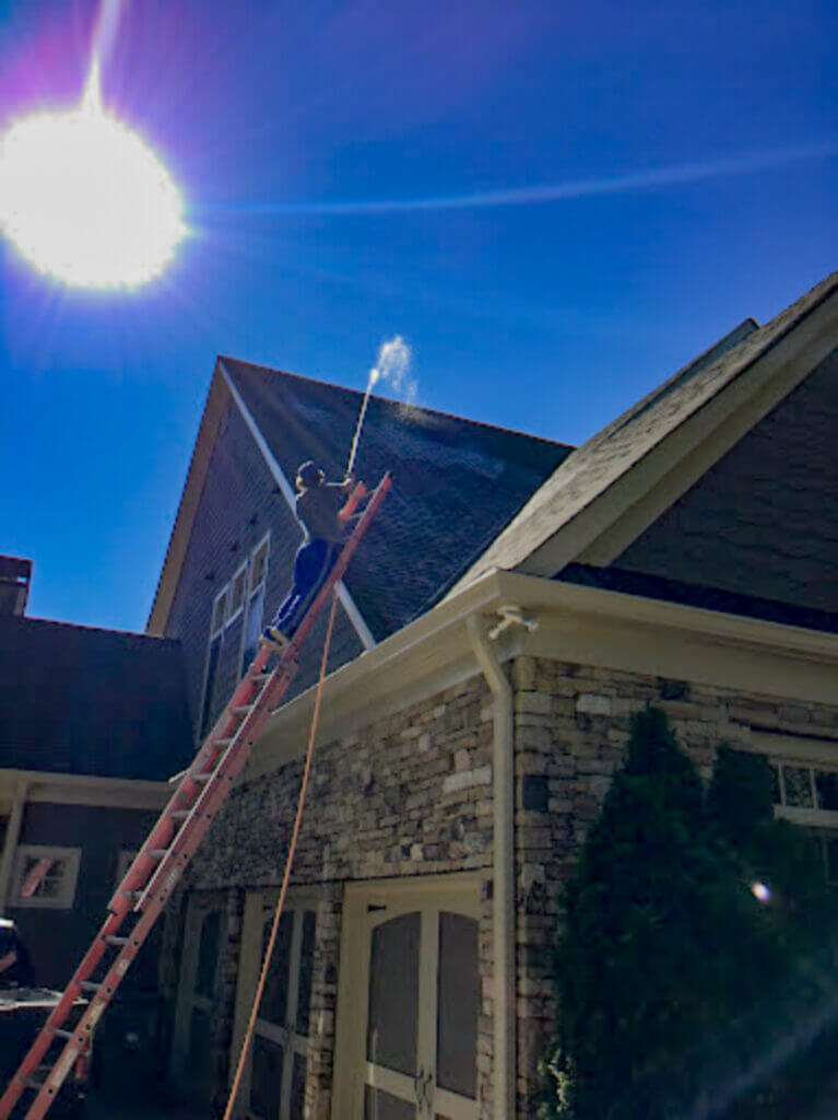 Blue Monkey tech on a ladder performing roof stain removal and pressure washing on a two-story home with eavestroughs and dormers.