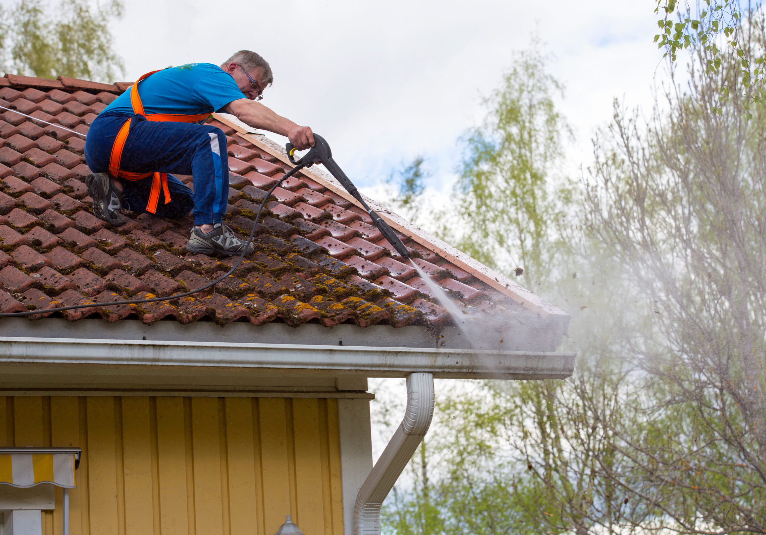 A Caucasian man wearing a safety harness pressure washes a roof and gutters on a slippery surface.