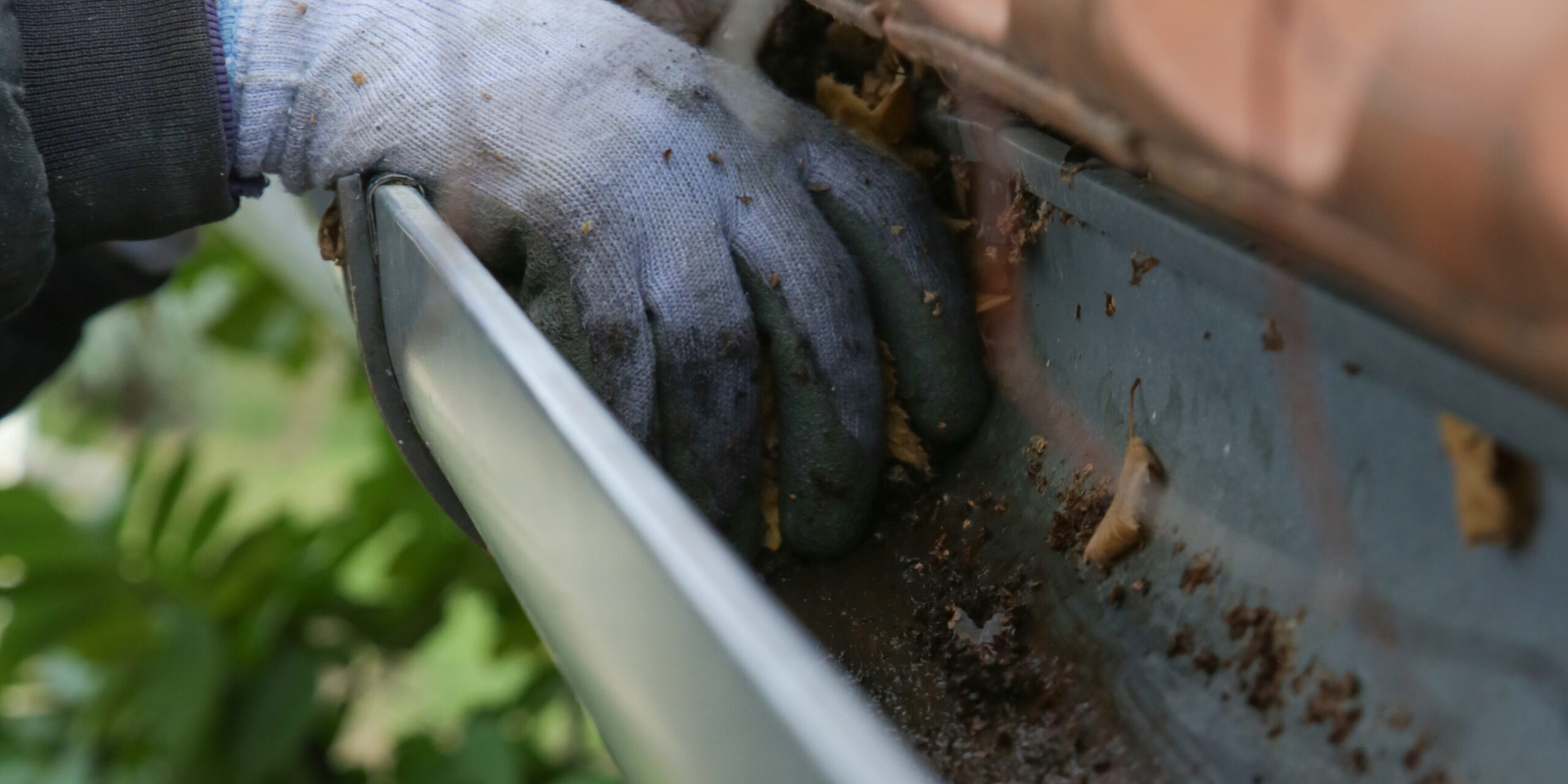 Expert Tips and Tricks for Easy Home Maintenance - Monkey Academy Blog Gloved hand removing leaves and debris from a clogged gutter during a professional gutter cleaning service.