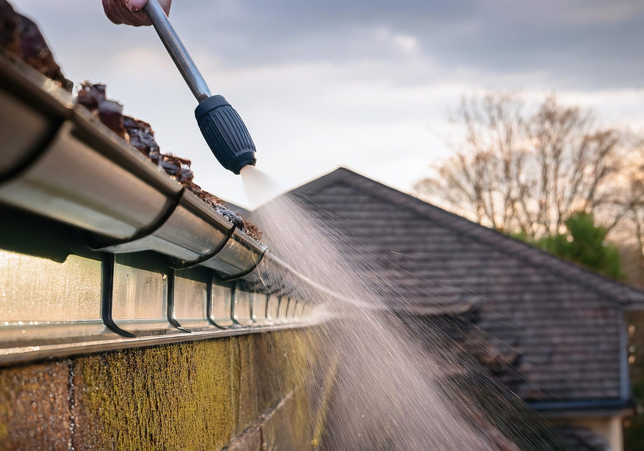 A SOFT-WASH PRESSURE WASHER REMOVES STAINS FROM THE EXTERIOR OF A GUTTER SURFACE - clears out leaf-filled spring rain gutters, removing seasonal debris.