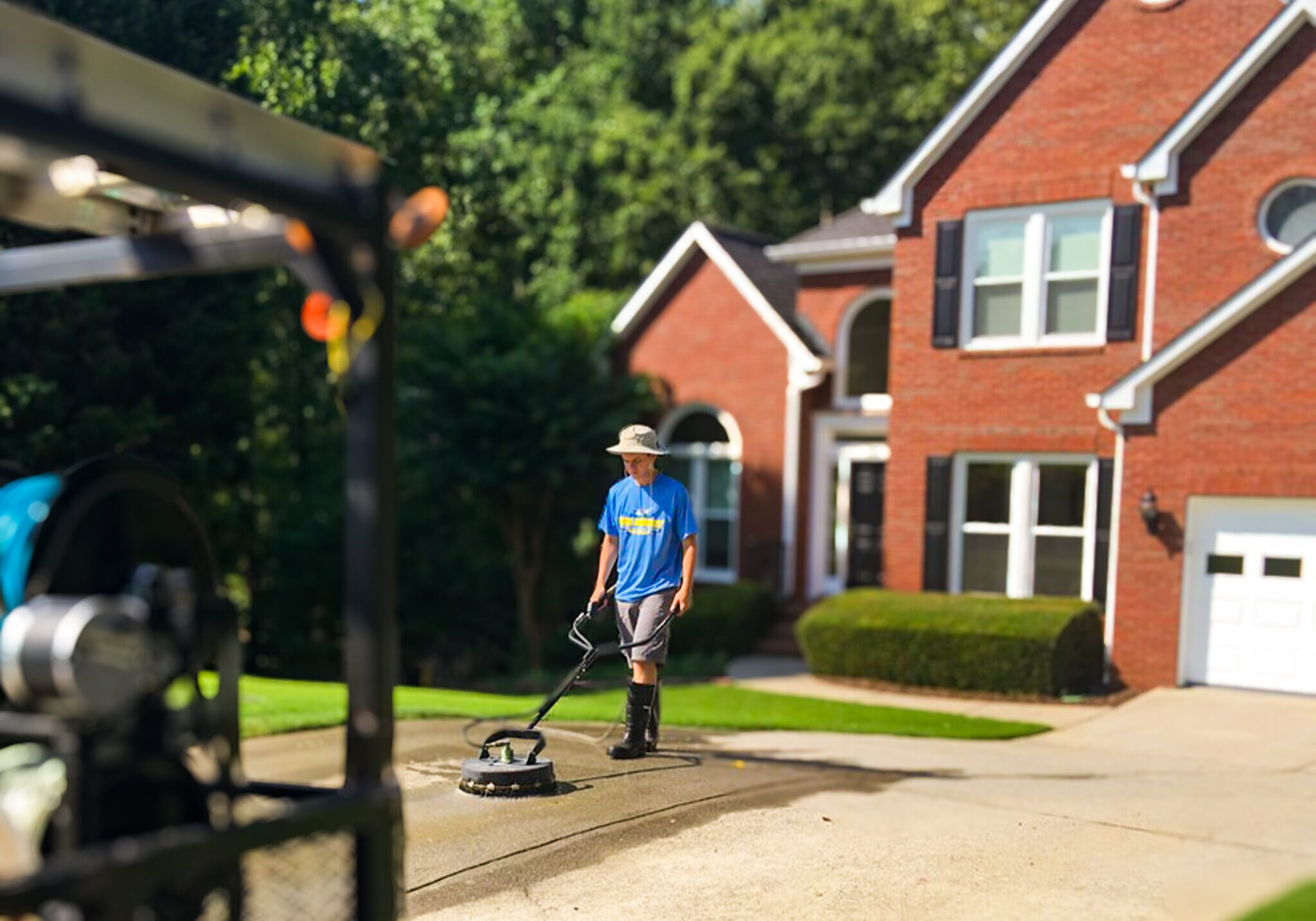Professional exterior cleaning service technician pressure washing a concrete driveway in front of a red brick home with a specialized surface cleaner.