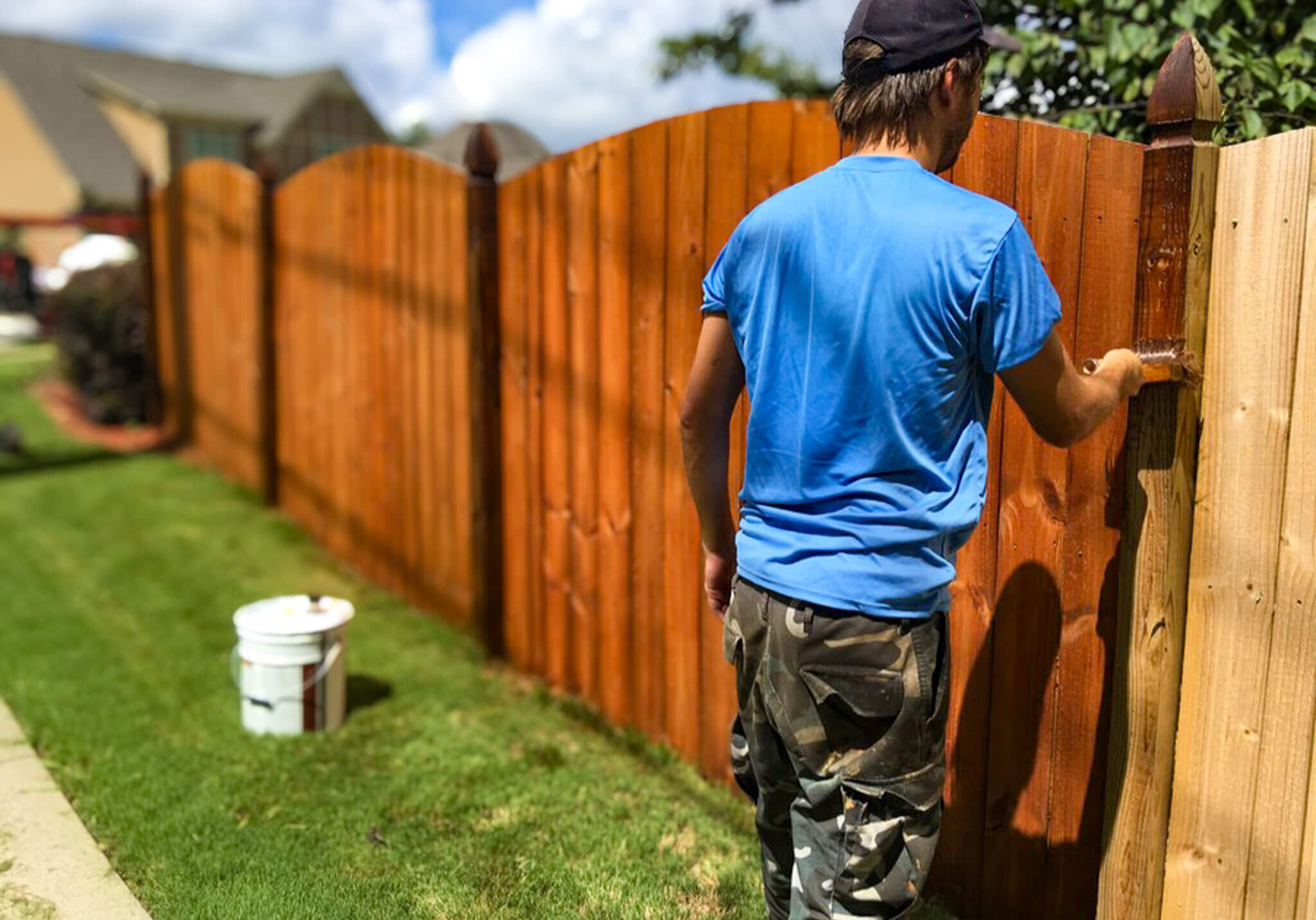 Man staining a wooden fence with a brush, applying a protective wood stain and sealant for long-lasting durability and rich color. FENCE PRESSURE WASHING. view our fence care services list