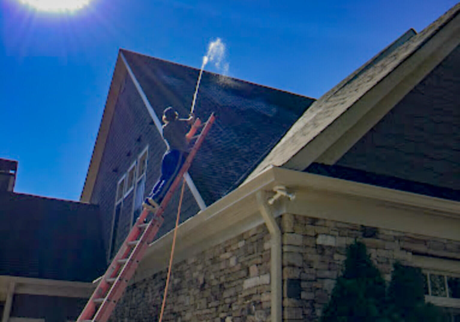 Blue Monkey tech on a ladder performing roof stain removal and pressure washing on a two-story home with eavestroughs and dormers.